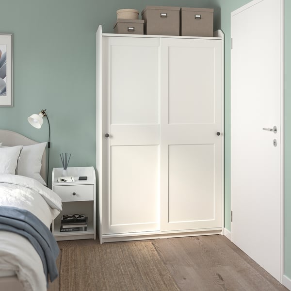 Modern bedroom with tall white HAUGA wardrobe, bed, nightstand, lamp, and books. Light blue walls, calming atmosphere.