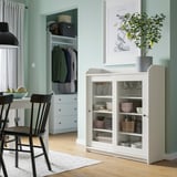 A white HAUGA glass-door cabinet, displaying dishes and bowls, positioned next to a dining area with black chairs and a plant on top.