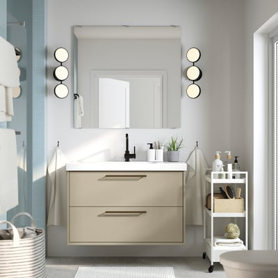Beige bathroom vanity with 2 drawers, wall-mounted. Includes ceramic sink, mirror, and lights. White storage cart with towels.