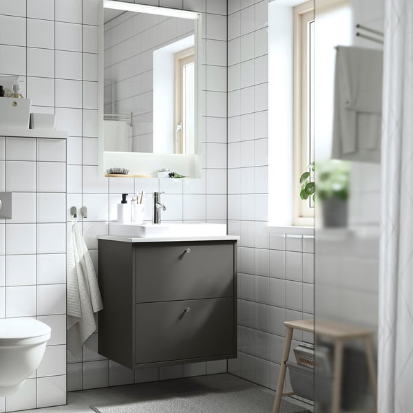 Modern bathroom with grey HAVBÄCK vanity, white sink, toilet, shower curtain, plant on stool, and white tiles.