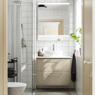 Modern bathroom with beige HAVBÄCK vanity, white basin, glass shower, mirror, and plant.