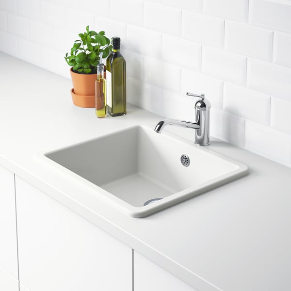 A modern white sink with a chrome faucet on a white counter. A plant and bottles beside it.