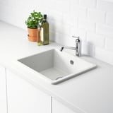 A modern white sink with a chrome faucet on a white counter. A plant and bottles beside it.