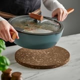 Person holding green casserole over cork stand on marble table.