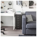 A HELMER black metal filing cabinet with five drawers on wheels beside a white desk and a grey armchair with a throw pillow.