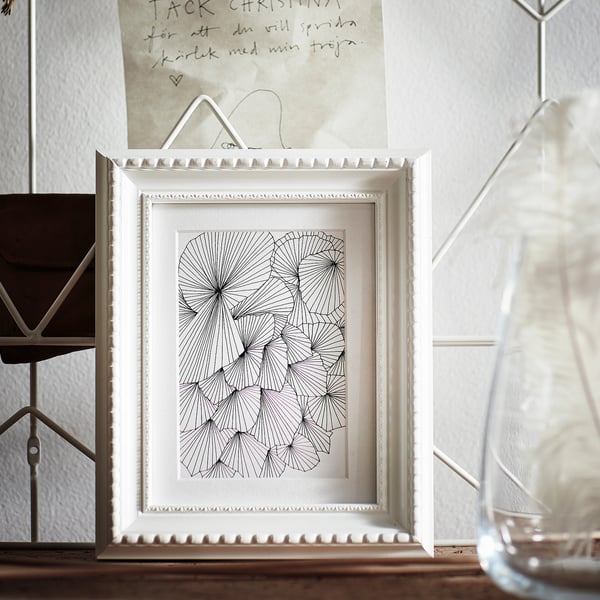 White framed geometric art print with intricate black designs, displayed on a wooden surface near a glass vase.