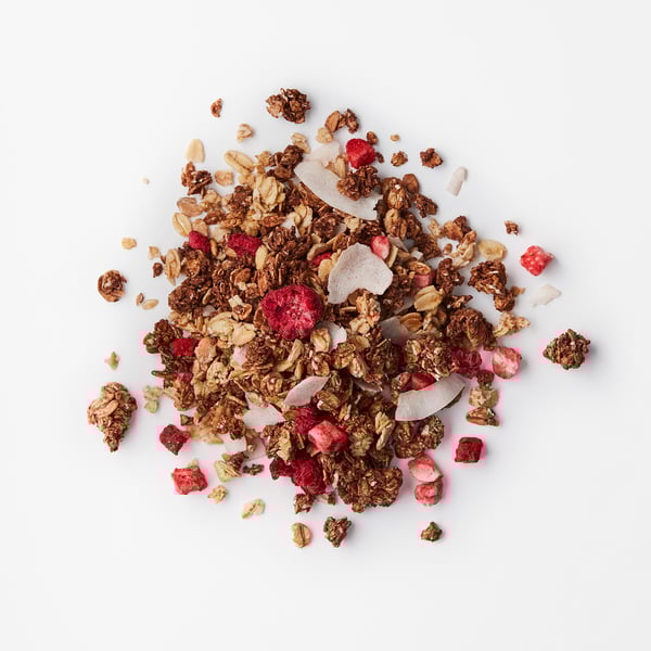 Heaped granola mix with crunchy chocolate oats, coconut flakes, and bright red dried berries on a white surface.