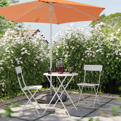 Small outdoor setup with orange umbrella, white table, and two chairs surrounded by blooming bushes.