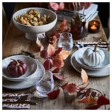 Wooden table set with white plates, glasses, HÖSTAGILLE autumn garland, small pumpkins. Cozy, warm decor with dark, rustic colors.