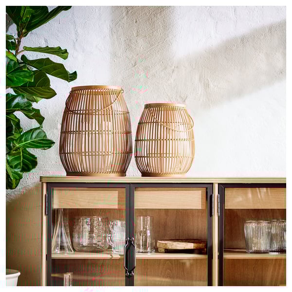 Bamboo lanterns with candles on wooden cabinet, large plant nearby, glassware inside.