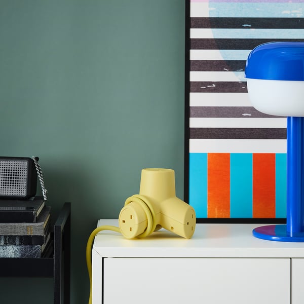 Bright yellow HORNFELS 4-way extension cord sits on a white drawer, smaller than a stack of books, near a blue lamp and green wall.