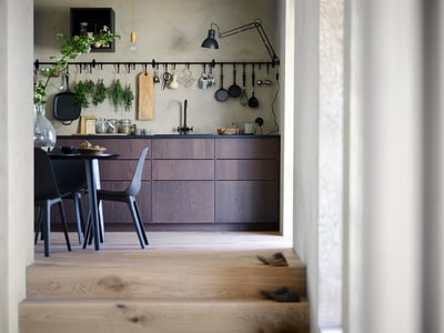 Modern kitchen with dark wood cabinets, hanging utensils, black chairs, and a dining table. Light wood floors and minimalist decor.