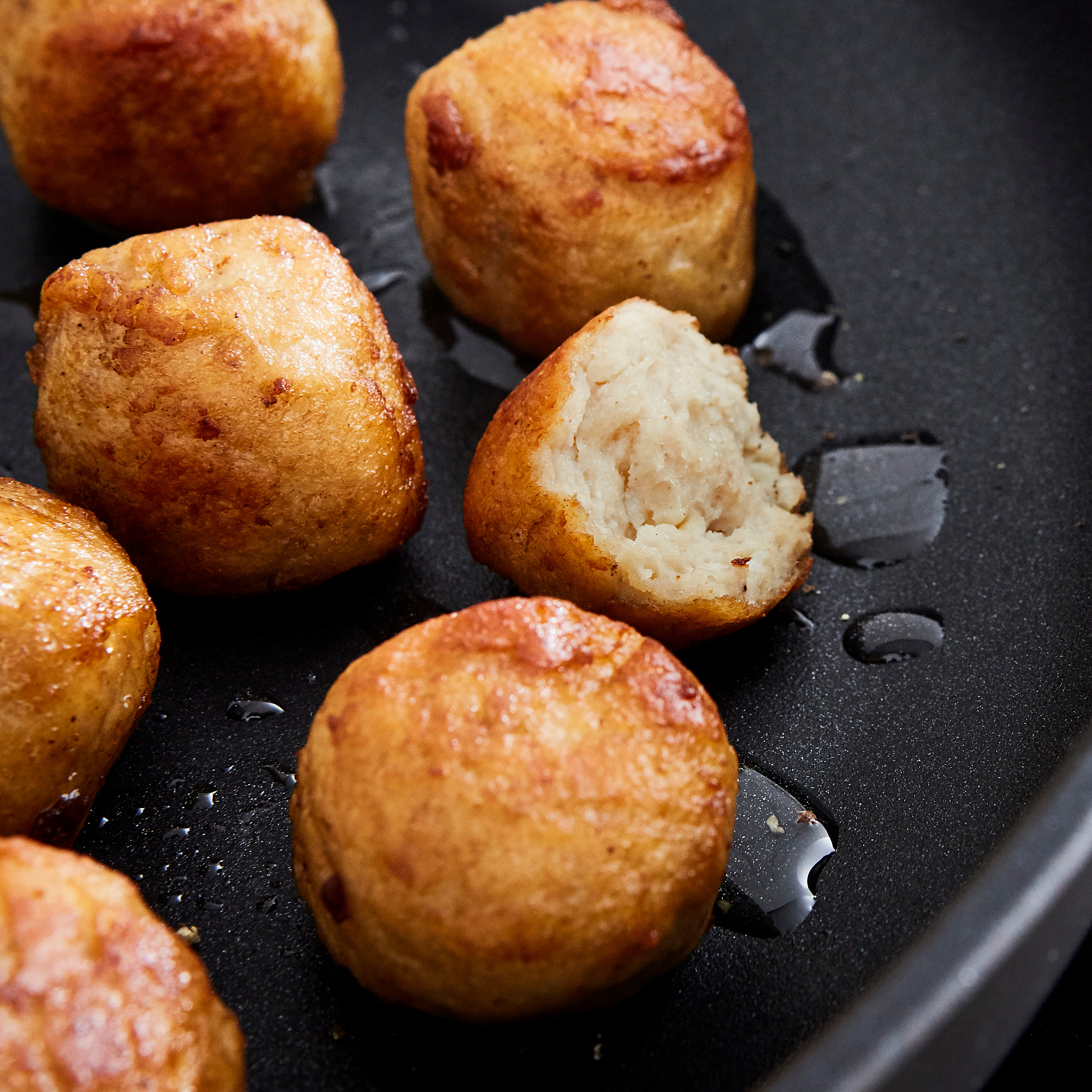 Golden-brown chicken meatballs cooking in a black skillet, one cut open revealing tender interior.