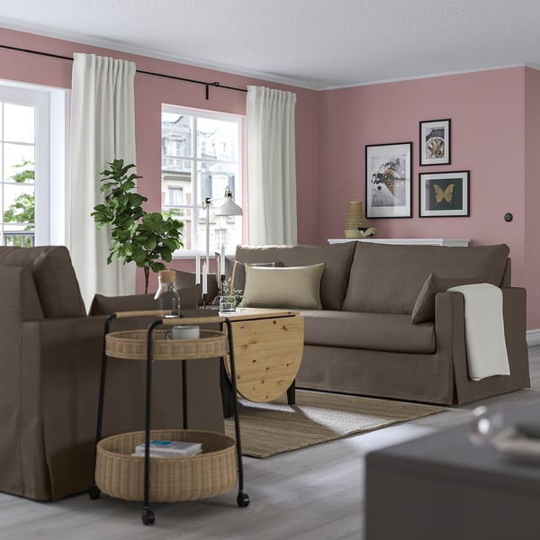 Cozy living room with HYLTARP sofa, window, armchair, and soft pink walls. Decor includes basket, tray, art, and blanket for comfort.