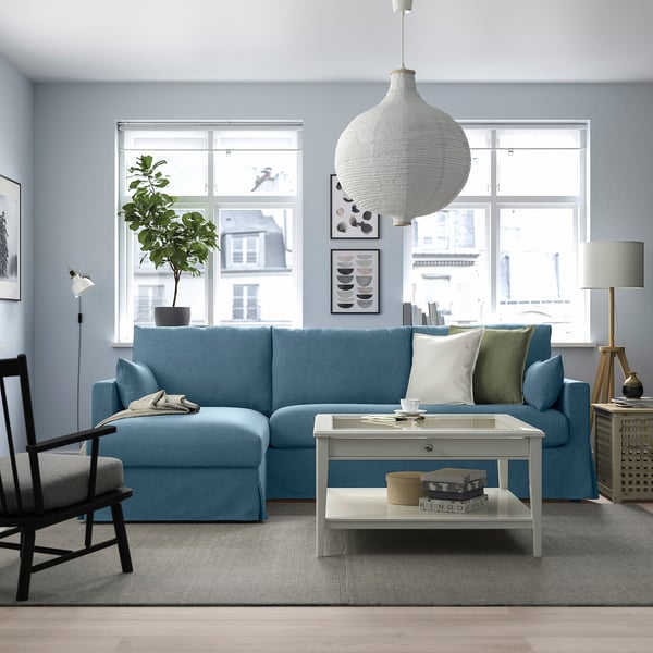 Modern living room with blue sectional sofa, city view window, white coffee table, wooden chair, tall lamp, pendant light, and houseplant.
