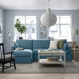 Modern living room with blue sectional sofa, city view window, white coffee table, wooden chair, tall lamp, pendant light, and houseplant.