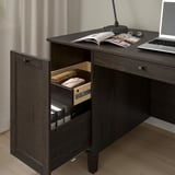Dark wood desk with open drawer revealing neatly stored items. Laptop and magazines on top, showcasing storage and workspace functionality.