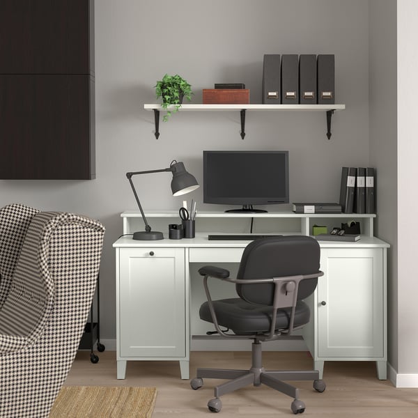 Modern home office setup with white IDANÄS desk, black chair, and monitor. Shelves hold books and a plant. Checkered armchair to side.