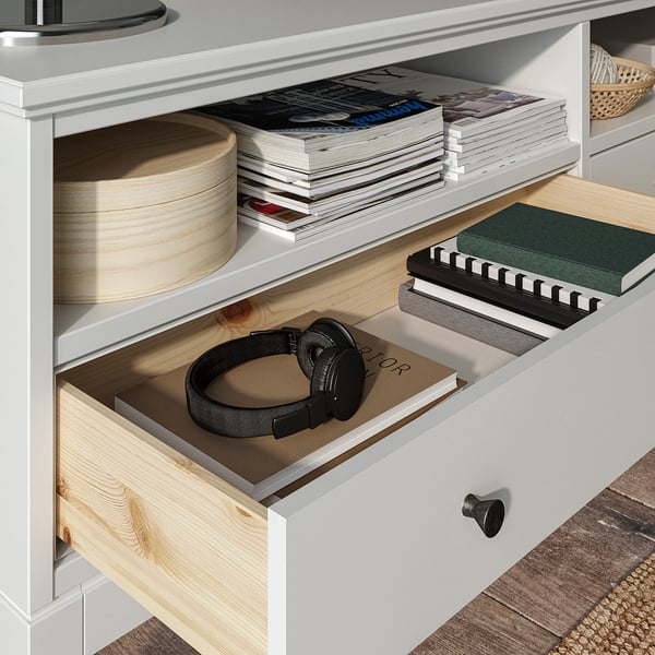 An IDANÄS white wooden storage unit with drawers and shelves. It contains books, headphones, and other items.