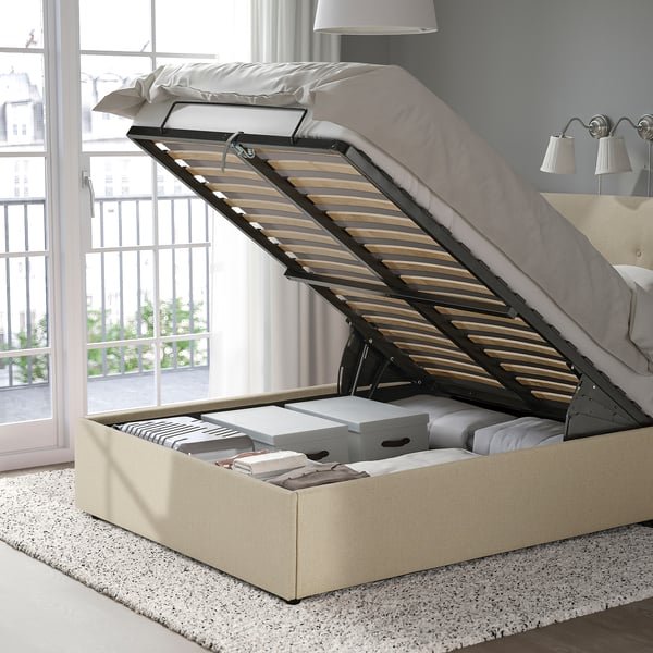 An upholstered bed with a lift-up mechanism, revealing storage space underneath. The bed has a sloping headboard and is placed near a large window.