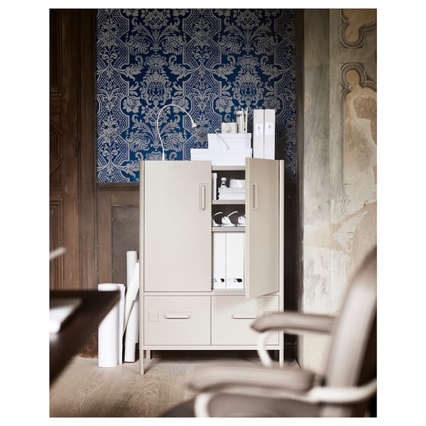 A white IDÅSEN office cabinet with a blue patterned wallpaper backdrop. The cabinet has open drawers and doors revealing storage items.