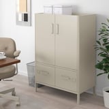 IDÅSEN beige metal cabinet in office setting. Two-door, two-drawer design with modern lines. Standing tall between chair and plant.