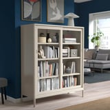 The IDÅSEN bookshelf in white with glass doors stands tall, holding books & decor. Its larger than the stool & sofa in a blue-walled room.