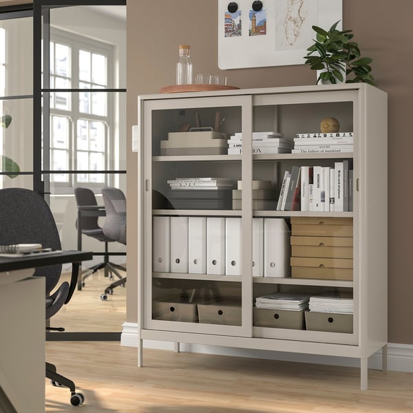 IDÅSEN white metal cabinet with glass doors in office, holding books & files, sleek design.