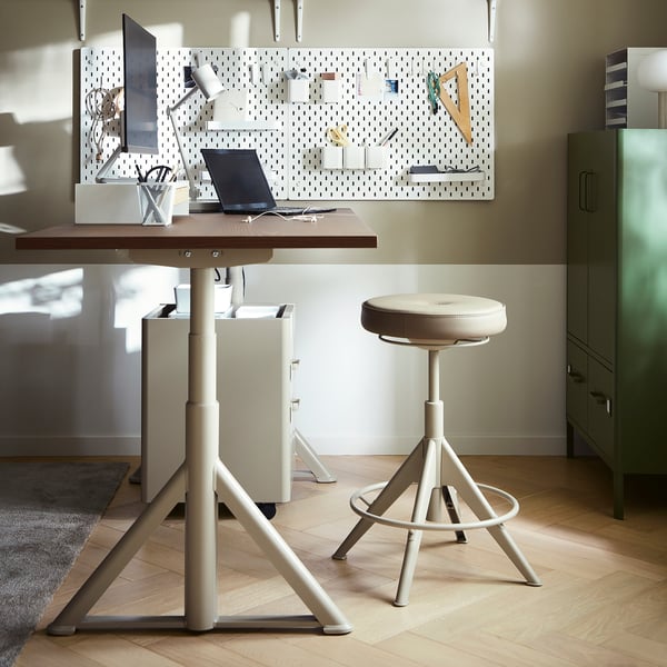 An IDÅSEN sit-stand desk in beige with a wooden top, a swivel chair, and a pegboard.