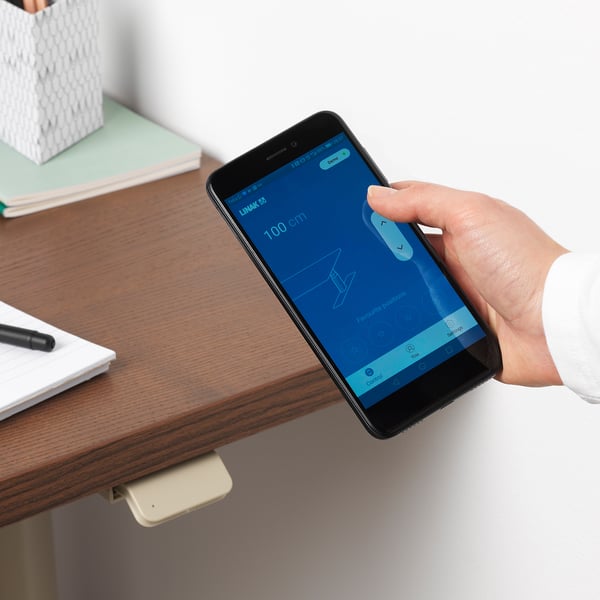 Hand holds smartphone app controlling height-adjusting desk, showing settings and adjustments.