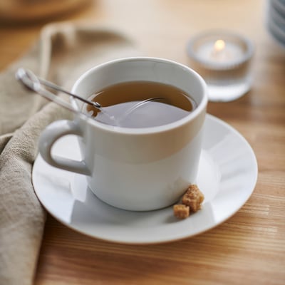 Cup of tea on white porcelain, with a spoon and sugar cube, placed on a wooden table.