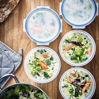 Six round food containers, four with lids on, filled with creamy vegetable soup, stacked and ready for storage or transport.
