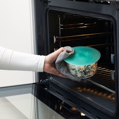 Hand places glass food container with green lid into oven. IKEA 365+ vacuum-sealing lid keeps leftovers fresh.