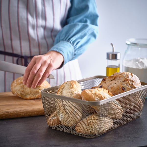 A person reaches into a mesh IKEA 365+ storage basket filled with loaves of bread, showcasing its breathable design for food storage.