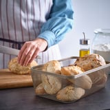 A person reaches into a mesh IKEA 365+ storage basket filled with loaves of bread, showcasing its breathable design for food storage.