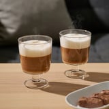 Two steaming coffee drinks in wide glass cups on a wooden table. Beside them, a bowl of chocolate treats.