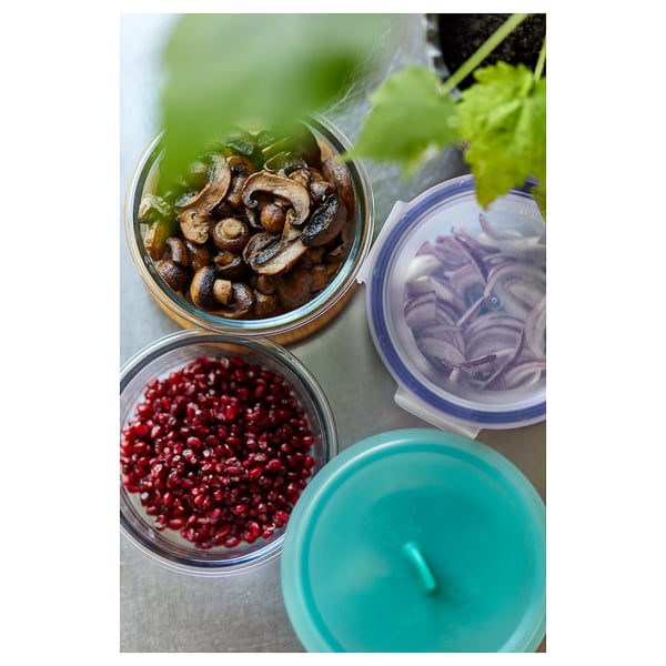 Three glass containers with blue, green, and clear lids hold mushrooms, pomegranate seeds, and sliced red onions, placed on a gray countertop.