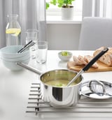 Modern kitchen setup with saucepan of soup on trivet, bowls, glasses, carafe, chair, and plant by window.