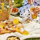 Picnic scene with orange drink ISKUB and snacks on a blanket. Person holds a bottle of the drink.