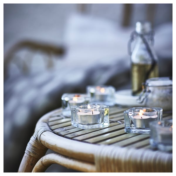 A set of glass candle holders, square and clear, on a wicker table, holding lit tealight candles.