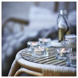 A set of glass candle holders, square and clear, on a wicker table, holding lit tealight candles.