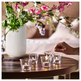 A hand lights clear glass candles on a wooden table beside a white vase with colorful flowers.
