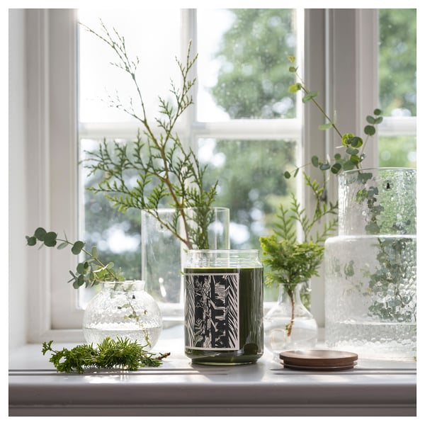 Dali: window sill with three vases. One round & clear, one green glass candle, and one round textured vase with greenery.