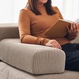 A woman reads a book on a beige JÄTTEBO sofa, wearing an orange top. The sofa a ripple-textured design for comfort and style.
