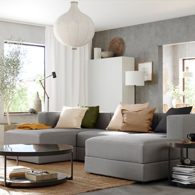Modern living room with grey JÄTTEBO modular sofa, colorful pillows, glass coffee table, and hanging lamp.