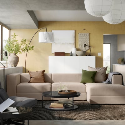 Modern living room with beige JÄTTEBO modular sofa, green pillows, black coffee table, large plant, and abstract art on mustard walls.