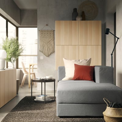 Modern minimalist living room: grey JÄTTEBO sofa with red pillows, wooden cabinet, macramé wall hanging, woven rug, and floor lamp.