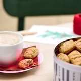 Image a white mug of frothy coffee on a red napkin, with cookies on a plate, and a tin of cookies beside it.