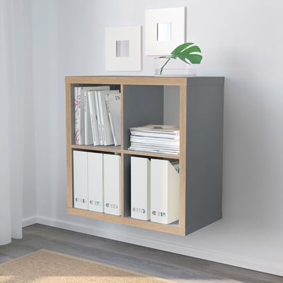 Gray KALLAX shelf with books, binders, plant, and empty frames in minimalist room.