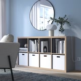 KALLAX bookshelf in light grey with light wood, rectangular, minimalist design. Sits low, fits beneath a round mirror, holding books and a lamp.